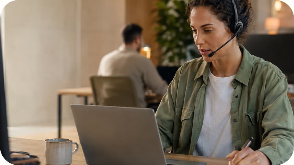 A woman wearing a headset works at a laptop in an office